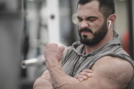 Muscle pain injury concept of young man with beard wearing wireless headset and hoodie holding hid damage biceps during hard workout in gymの写真素材