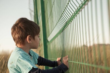 poor dirty little caucasian boy refugee standing near state border fence holding it with hands with hope of help and seeking for refugeの写真素材