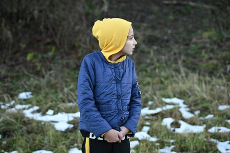 frozen little girl in yellow hoodie and blue jacket shiver near snow on ground in outdoor park during leisure hikingの写真素材
