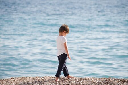 one lonely preschool kid in white jersey and blue trousers walking upset along sea beach in springの写真素材