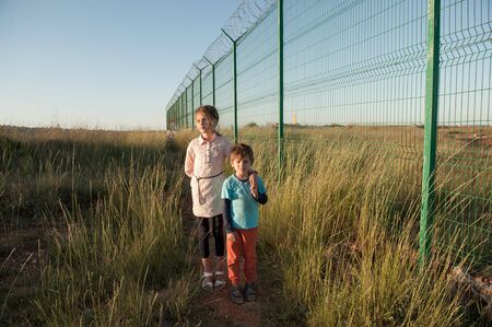 two poor kids brother and sister refugees near state border with long fence with barbed wireの写真素材