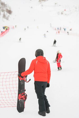 man in black helmet wearing winter sportswear holding snowboard looking at snow mountain with skiers and snowboarder young girl in red clothesの写真素材