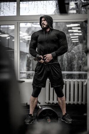 mental and physical growth concept of hard workout training in gym by strong young athlete man wearing hoodie and powerlifting belt near barbellの写真素材