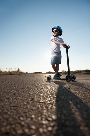 active sport little child in blue helmet and shorts wearing trendy sunglasses standing on asphalt road with mini scooter on hot sunset summer dayの写真素材