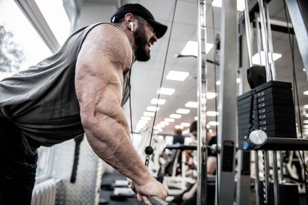 powerful strong man with beard wearing cap and sportswear pumping iron in sport gym for muscle development listening music using wireless headphonesの写真素材