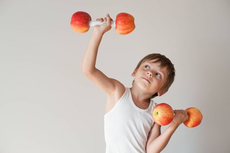 home leisure activity of young small kid in white tank top lifting dumbbells made of red applesの写真素材
