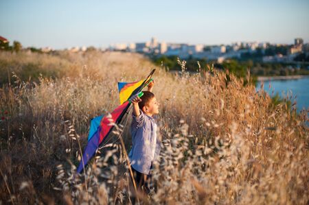 happy little boy with colorful kite standing among yellow high grass on sunset near city with port sea bayの写真素材
