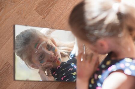 beautiful little child girl with face painted with color tint in dress looking at her reflection in mirror on floorの写真素材