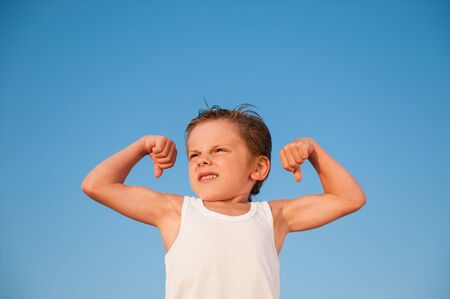 strong little boy in white tank top showing strong muscle on blue sky backgroundの写真素材