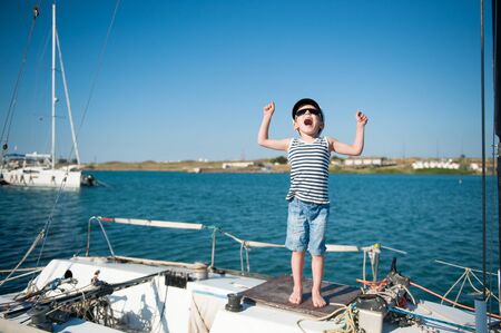 happy active small kid in sailor hat shouting with joy on white yacht board during summer travel vacationの写真素材