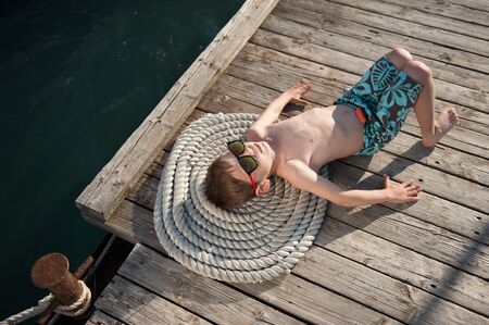 relaxation of healthy idle little child in sunglasses and trendy shorts lying dreaming on wood pier sea waterの写真素材