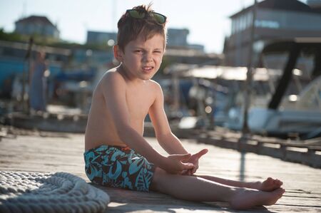 funny healthy cute little boy in shorts sitting on wooden sea pier on sunny summer day squintingの写真素材