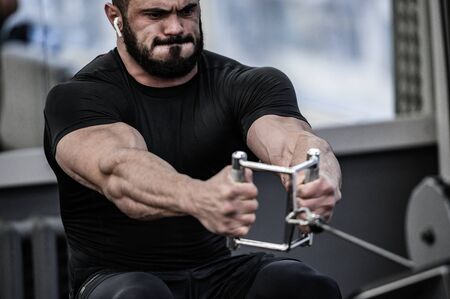 strong young bearded caucasian male in black jersey wearing wireless headphones lifting heavy weight in sport club with grimace of stress on his face overcoming painの写真素材