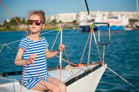 graceful elegant small girl in sunglasses and striped shirt sitting on board of sea white yacht on summer travel vacationの写真素材