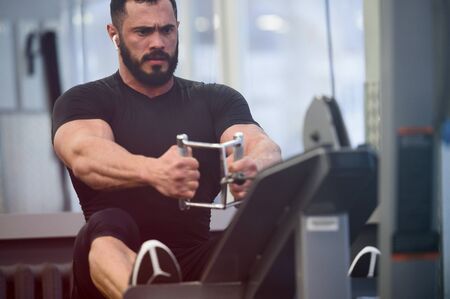 male health concept of heavy weight training of young handsome man with beard making exercise on sport fitness simulator equipmentの写真素材