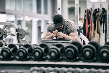 heavy stress training concept of young bearded tired male in black cap resting after hard workout in fitness indoor gym among iron dumbbellsの写真素材