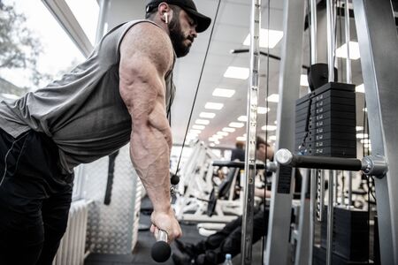 powerful strong athlete caucasian man with beard wearing cap and sportswear pumping iron in sport gym for muscle development listening music using wireless headphonesの写真素材