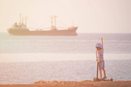 happy little child in helmet with scooter on sea beach with cargo ship on horizon on sunsetの写真素材