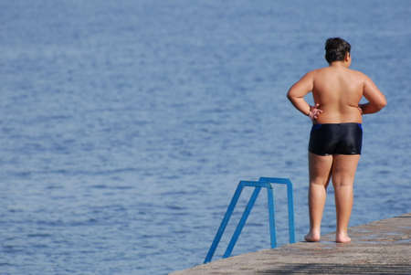 overweight obesity concept of one little thick fat boy in swimming shorts standing on beach during summer vacationの写真素材