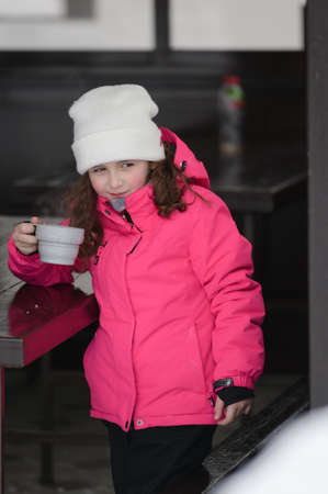 cute little girl in ski suit drinking hot tea from cup during cold winter outdoor leisure activity on mountain resortの写真素材