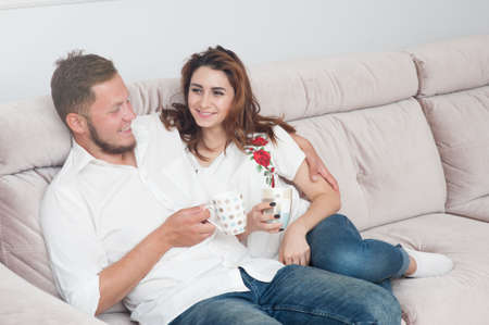 happy young couple caucasian man and woman sitting at sofa with cup of tea at homeの写真素材