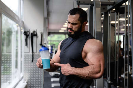handsome strong young athlete male with beard holding bottle shaker with blue energy drink with copy space in sport gymの写真素材