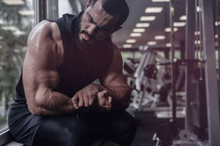 strong young bearded man in black clothes showing muscles biceps sitting near window in sport gym with exercise equipment and copy spaceの写真素材