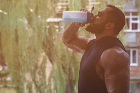 strong young athlete drinking energy drink during from bottle shaker heavy training workout in gym with big window and copy spaceの写真素材