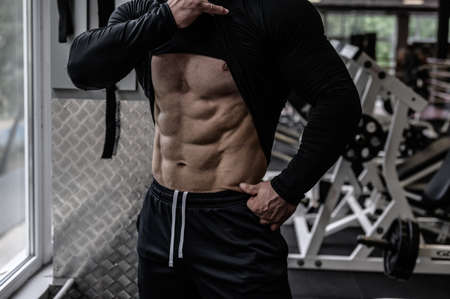 perfect body with abdominal muscles shown by young strong athlete man near window in sport gym with exercise equipmentの写真素材