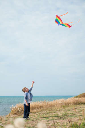 one active healthy small kid holding flying in air colorful kite standing on sea shore with blue sky on sunny summer holidayの写真素材