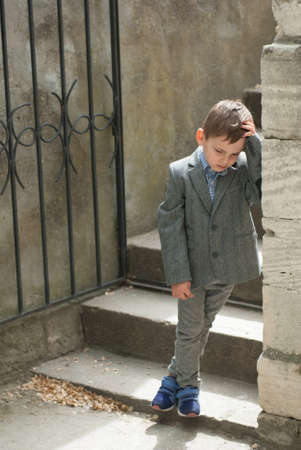 Sad little boy on the background of an old wall and stairs with a gateの写真素材