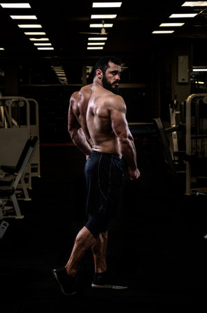 strong young bearded caucasian man with sport physique body standing in dark fitness gym showing powerful back muscleの写真素材