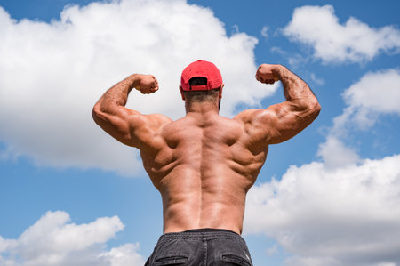 strong young athlete fitness bodybuilder male in red cap showing his powerful muscular back development on cloudy blue sky background with copy spaceの写真素材