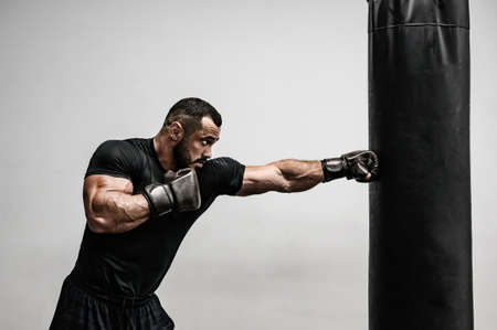 strong young caucasian bearded male in black sportswear kick punching bag with hands in boxing gloves on bright background in gym with copy spaceの写真素材