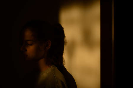 domestic violence abuse concept of little sad child girl standing in shadow lit with sunset light indoors at homeの写真素材