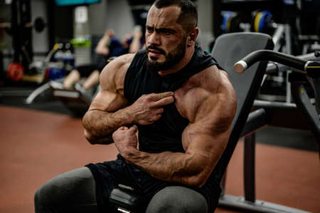 strong bearded caucasian man bodybuilder watching his shoulder and arm muscle sitting in sport fitness athletic gymの写真素材