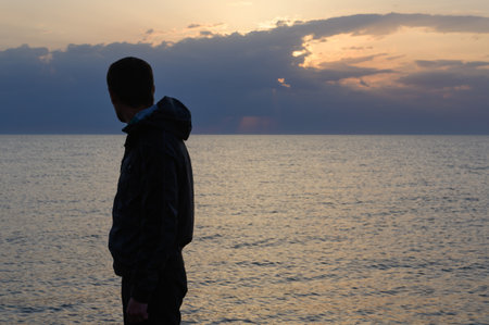 memories concept of silhouette of adult male in jacket with hood standing on sea shore looking at beautiful sky on ocean twilight duskの写真素材