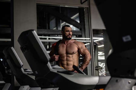 strong young bearded caucasian male with big muscles physique in sport fitness gym at night during relaxation after heavy workout training among treadmillの写真素材