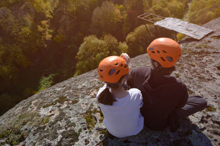 two young kids in orange helmet sitting on top of high rocky mountain with forest down below during autumn extreme hiking climbing leisure activityの写真素材