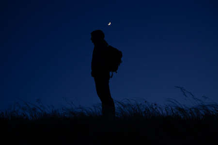 one lonely man tourist with backpack standing among windy field at night with moon shining above during travel hikingの写真素材