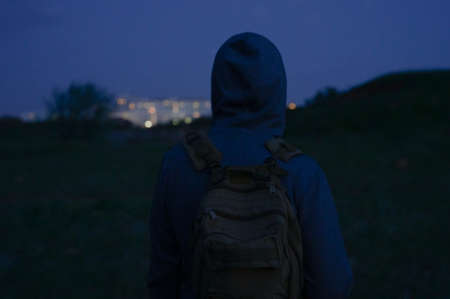 homeless man in hoodie with backpack watching on night city buildings during dusk hiking aloneの写真素材