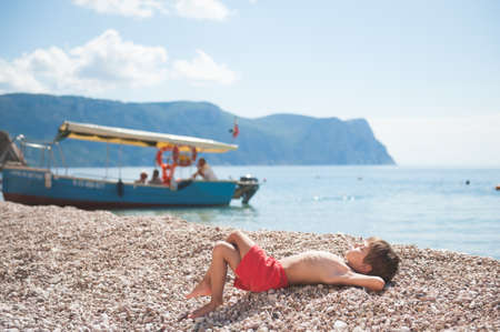 happy healthy dreaming little child lying on summer sea beach with boat and rocky mountain on backgroundの写真素材