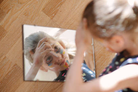 beautiful little girl with face painted with paint looking on her reflection in mirrorの写真素材