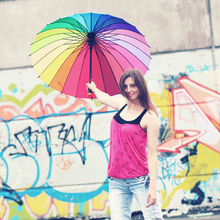 An ordinary girl with a rainbow umbrella on the roof of a house on a background of graffitiの写真素材
