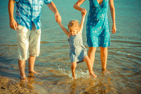 View of happy young family having fun on the beach. Young Mom and Dad dabbles with daughter in the water near the shore.の写真素材