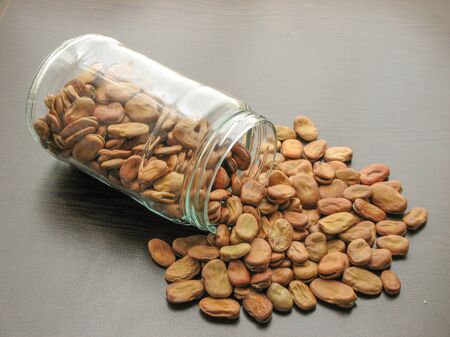 Broad beans (Vicia faba) are poured from a glass jar on brown wooden surface. Broad beans in a glass...