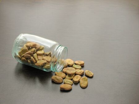 Broad beans (Vicia faba) are poured from a glass jar on brown wooden surface.  Broad beans (Vicia faba) in a glass jar on the wooden table. Broad beans (Vicia faba) in a glass dish. Wooden backgroundの写真素材