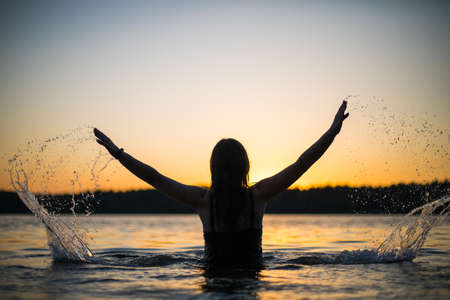 Beautiful Russian girl in a long black swimsuit swims outside the city on the lake in the rays of sunset or dawn. Splashing water on the background of the sun. Happy girl.の写真素材