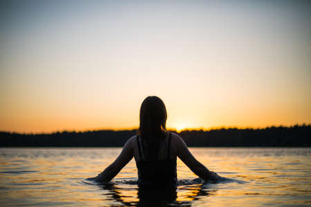 Beautiful Russian girl in a long black swimsuit swims outside the city on the lake in the rays of sunset or dawn. Splashing water on the background of the sun. Happy girl.の写真素材