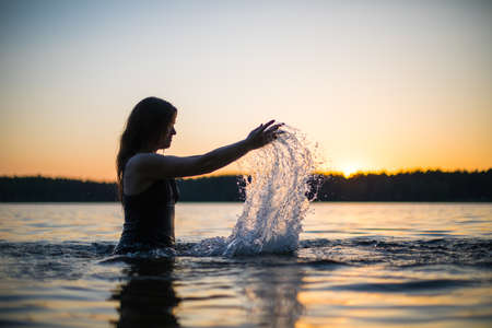 Beautiful Russian girl in a long black swimsuit swims outside the city on the lake in the rays of sunset or dawn. Splashing water on the background of the sun. Happy girl.の写真素材